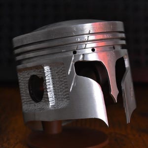 Peut inclure: Un piston de moteur métallique modifié en forme de casque avec une découpe de visière en T. L'objet argenté présente des rainures horizontales sur le dessus, un panneau latéral texturé et une section avant lisse et incurvée rappelant un masque.