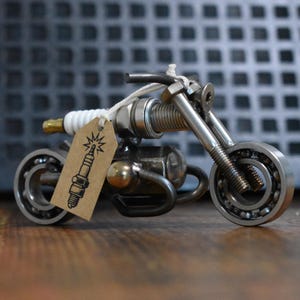 Op de afbeelding: Een miniatuur motorfietssculptuur gemaakt van gerecyclede metalen auto-onderdelen, waaronder kogellagers voor de wielen en een bougie voor het lichaam. Een klein bruin kartonnen label met een zwarte inkttekening van een bougie is bevestigd.