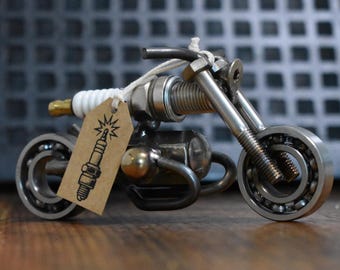 May include: A miniature motorcycle sculpture crafted from recycled metal automotive parts, including ball bearings for wheels and a spark plug for the body. A small brown cardboard tag with a black ink illustration of a spark plug is attached.