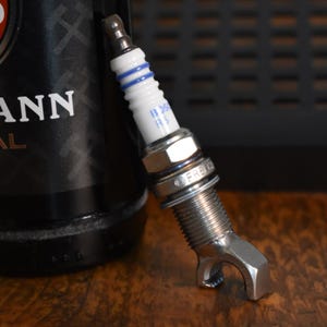Könnte beinhalten: Eine Zündkerze aus Metall mit weißem Keramikisolator, zwei blauen Ringen und dem Schriftzug BOSCH. Der untere Teil der Zündkerze ist zu einem Flaschenöffner umgeformt, mit einem metallischen Gewindekörper und einer dunklen Metallspitze.