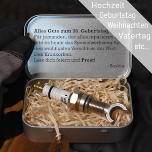 May include: A silver metal tin containing a spark plug modified into a bottle opener, nestled in wood shavings. The lid interior features German text: Alles Gute zum 30. Geburtstag, Für jemanden, der alles reparieren kann, gibt es heute das Spezialwerkzeug.