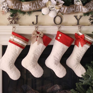 May include: Four white quilted Christmas stockings with red and brown trim hanging on a fireplace mantel. The stockings have different embellishments, including a bow, buttons, and stripes. The word "JOY" is displayed above the stockings.