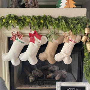 May include: Four Christmas stockings hang above a fireplace. Each stocking is a different color and texture, with a bow at the top and a name embroidered on the front: Boomer, Dixie, Zero, and Kevin. Green garland and lights decorate the mantel.