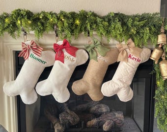 May include: Four Christmas stockings hang above a fireplace. Each stocking is a different color and texture, with a bow at the top and a name embroidered on the front: Boomer, Dixie, Zero, and Kevin. Green garland and lights decorate the mantel.