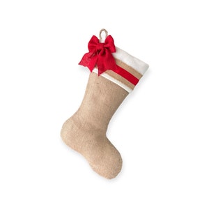 May include: A burlap Christmas stocking with a white cuff and a red bow. The stocking has a red and white striped ribbon around the cuff.
