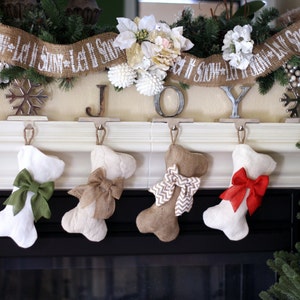 May include: A festive Christmas mantelpiece with four bone-shaped stockings, each adorned with a different colored bow. The mantel is decorated with a burlap banner that reads "Let It Snow" and the word "JOY" in individual letters.