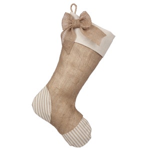 May include: A brown burlap Christmas stocking with a white cuff and a brown burlap bow. The stocking has a white and brown striped pattern on the bottom.