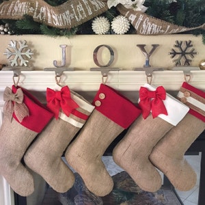 Op de afbeelding: Vijf kerstsokken van jute met rode bovenkanten en decoratieve strikken of knopen hangen aan een schoorsteenmantel. Het woord "JOY" staat met zilveren letters boven de sokken. De schoorsteenmantel is versierd met groen en sneeuwvlokken.