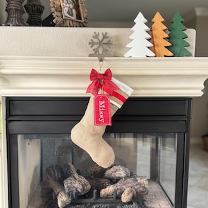 May include: A tan hessian Christmas stocking featuring a white cuff with two red stripes, a red bow, and a red rectangular tag with the name Missy embroidered in white, hanging from a silver snowflake-shaped metal stocking holder.
