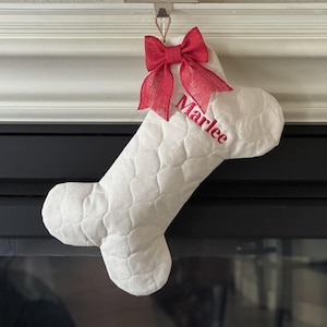 May include: A white, bone-shaped Christmas stocking with a quilted pattern. The stocking has a red burlap bow at the top and the name "Marlee" embroidered in red. It hangs from a metal hook.