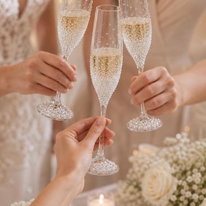 May include: Three tall, clear glass champagne flutes held in a toast. Each flute features a slender stem and a bowl decorated with numerous small, white, spherical beads that cluster densely toward the base and scatter upward.