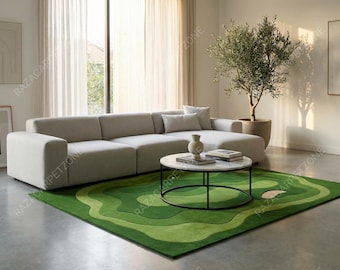 May include: A modern living room featuring a large, light gray sectional sofa, a round coffee table with a marble top, and a green rug with a golf course design. A potted olive tree adds a natural element.
