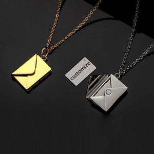 Könnte beinhalten: Zwei Umschlag-Halsketten, eine goldfarbene und eine silberfarbene, mit passenden Ketten. Ein kleines weißes Etikett mit dem Wort "customize" ist ebenfalls vorhanden. Die Halsketten befinden sich auf einer schwarzen Oberfläche.