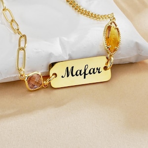 Könnte beinhalten: Eine goldfarbene Halskette mit einem rechteckigen Anhänger, auf dem der Name "Mafar" eingraviert ist. Die Kette hat eine Kette mit einem quadratischen rosa Edelstein und einem ovalen gelben Edelstein. Der Anhänger ist 3 cm lang.
