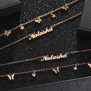Könnte beinhalten: Gold- und roségoldfarbene Halsketten mit dem Namen "Natasha" in Schreibschrift. Die Ketten sind mit Schmetterlings- und Sternanhängern verziert. Der Hintergrund ist dunkel und glitzernd.
