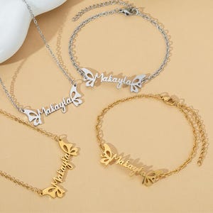 Könnte beinhalten: Eine Sammlung personalisierter Schmuckstücke, darunter Halsketten und Armbänder. Jedes Stück trägt den Namen "Makayla" in zarter Schrift, akzentuiert durch schmetterlingsförmige Verzierungen. Der Schmuck ist in Silber- und Goldtönen erhältlich.