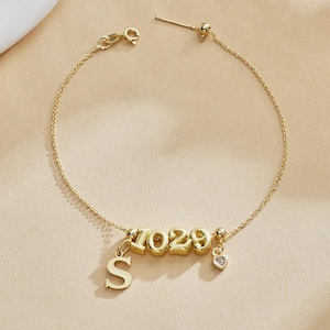 Könnte beinhalten: Ein goldfarbenes Armband mit den Zahlen "1029" und dem Buchstaben "S" als Anhänger. Ein kleiner herzförmiger Anhänger mit einem klaren Stein hängt am Armband. Das Armband befindet sich auf einem neutralen Hintergrund.