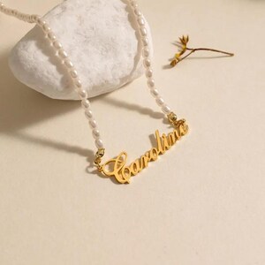 Könnte beinhalten: Eine zarte Perlenkette mit einem goldenen Anhänger mit dem Namen "Caroline" in Schreibschrift. Die Kette besteht aus kleinen, ovalen Perlen. Der goldene Anhänger verleiht der Kette einen Hauch von Eleganz.