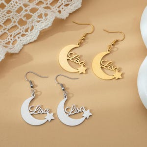 Könnte beinhalten: Gold- und silberfarbene Ohrringe mit einem Halbmond-Design und dem Namen "Lisa" in Schreibschrift. Jeder Ohrring hat einen kleinen Sternanhänger. Die Ohrringe werden auf einer hellbraunen Oberfläche präsentiert.