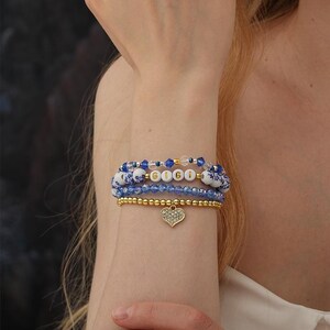 Könnte beinhalten: Mehrere Armbänder in Blau- und Goldtönen. Eines der Armbänder zeigt den Namen "GIGI" mit weißen Buchstabenperlen. Ein goldener Herzanhänger mit klaren Steinen hängt an einem Armband. Die Armbänder werden am Handgelenk getragen.