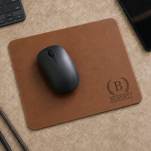 Peut inclure: Un tapis de souris rectangulaire en cuir marron avec une souris d'ordinateur sans fil noire posée dessus. Le tapis de souris est gravé d'un motif de couronne de laurier entourant la lettre B et le nom BENNETT écrit en lettres capitales.