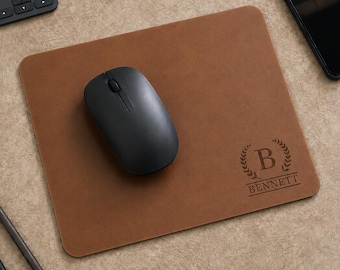 May include: A rectangular brown leather mouse pad featuring a black wireless computer mouse placed on its surface. The mouse pad is engraved with a laurel wreath design surrounding the letter B and the name BENNETT printed in capital letters.