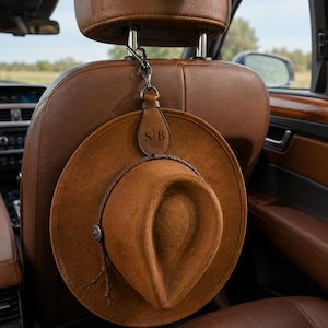 May include: A brown felt hat with a braided band and a metallic side accent, suspended from a car headrest by a brown leather strap featuring the embossed letters SB. The hat is centred against the back of a brown leather car seat.
