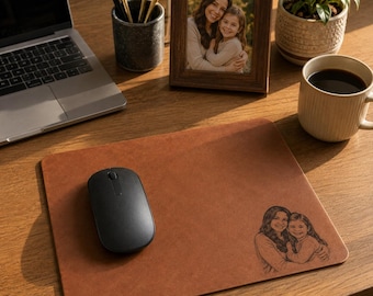 May include: A rectangular tan leather mouse pad featuring a laser-engraved illustration of a woman and a young child embracing. The mouse pad is positioned on a wooden desk surface next to a black wireless computer mouse.