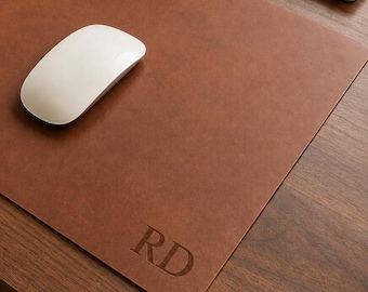 May include: A rectangular brown desk mat with a smooth surface and rounded corners. The mat features the embossed letters RD in the bottom right corner. A white wireless computer mouse is positioned on the left side of the brown desk mat.