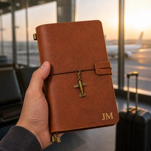 Può includere: Un diario in pelle marrone con chiusura a fascia elastica verticale e un ciondolo a forma di aereo color bronzo. La copertina presenta le iniziali JM impresse nell'angolo in basso a destra e due piccoli terminali metallici color bronzo.