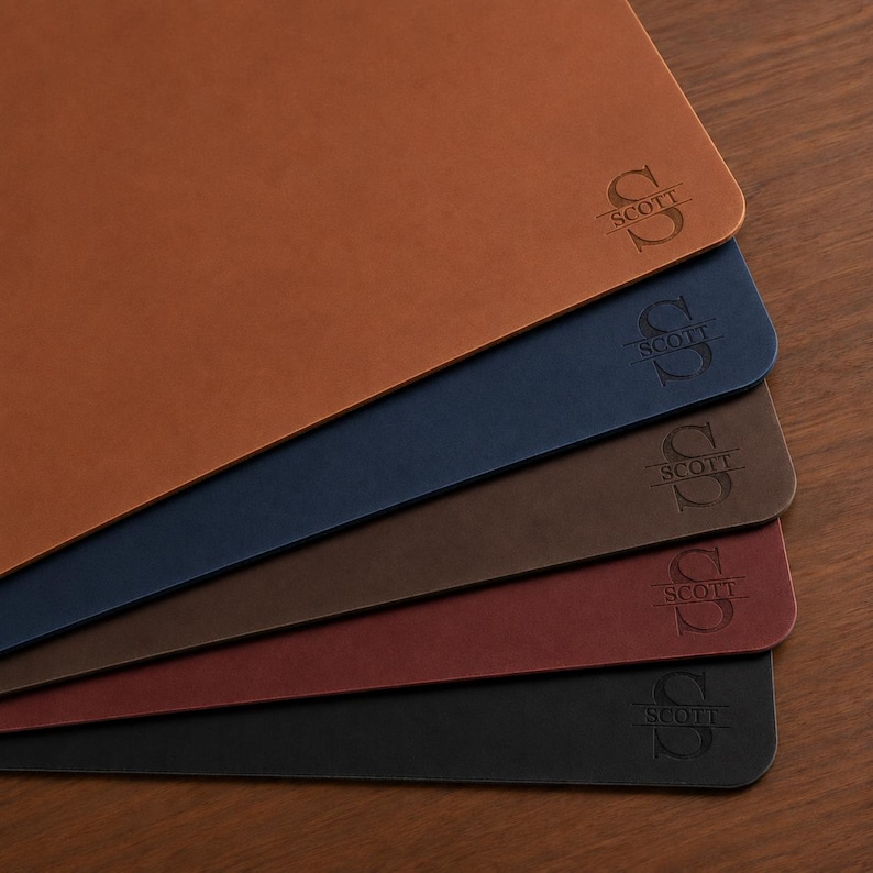 May include: A stack of five rectangular leather desk mats in tan, navy blue, dark brown, burgundy, and black. Each mat features a debossed monogram design consisting of a large letter S with the name SCOTT centered horizontally across the letter.
