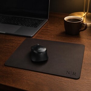 May include: A rectangular dark brown leather mouse pad featuring the embossed letters N | R in the bottom right corner. A black Logitech computer mouse is positioned in the center of the smooth surface, which is placed on a wooden desk.