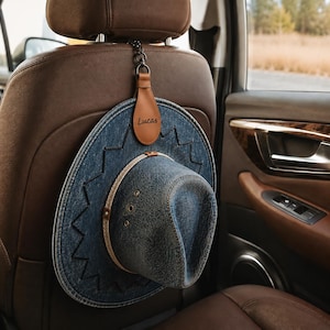 Puede incluir: Un sombrero vaquero de mezclilla cuelga del reposacabezas de un asiento de coche con una correa y cadena de cuero marrón. La correa está grabada con el nombre "Lucas". El sombrero tiene un ala clara y costuras decorativas.