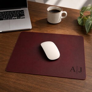 May include: A rectangular dark burgundy leather mouse pad featuring the embossed initials A and J separated by a vertical line in the bottom right corner. The mouse pad has a smooth surface and rounded corners, with a white computer mouse placed on top.