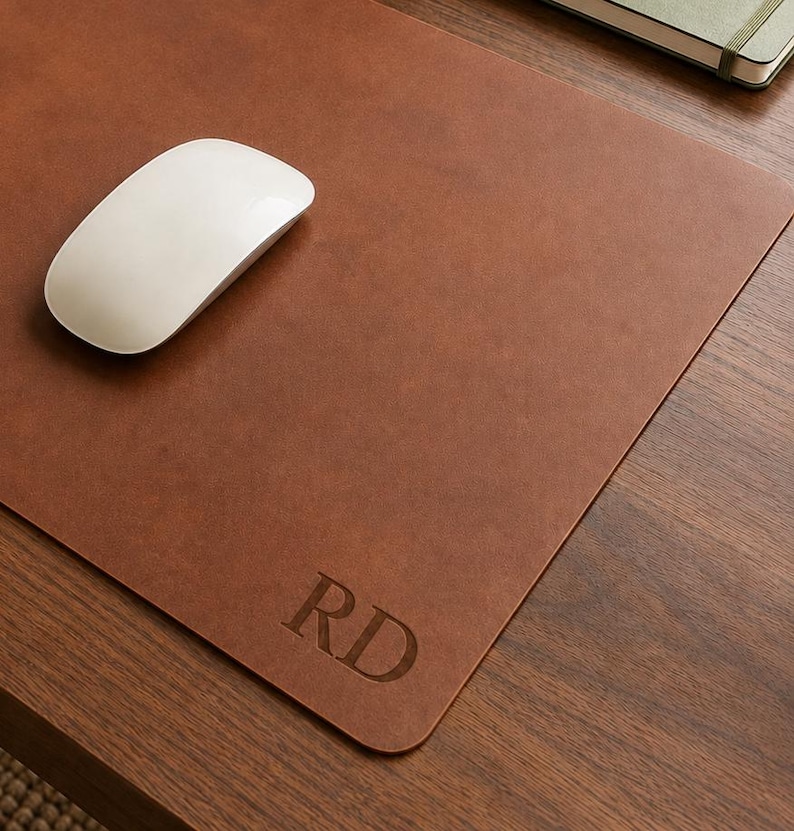 May include: A rectangular brown desk mat with a smooth surface and rounded corners. The mat features the embossed letters RD in the bottom right corner. A white wireless computer mouse is positioned on the left side of the brown desk mat.