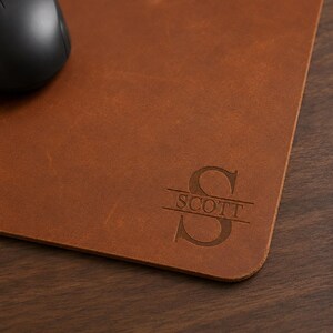 May include: A rectangular brown leather mouse pad featuring a debossed monogram design. The design consists of a large capital letter S with the name SCOTT engraved horizontally across the center of the letter. A black computer mouse is visible.