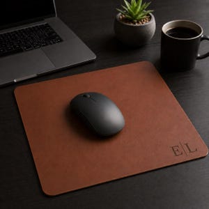 May include: A square brown leather mouse pad featuring the embossed letters E and L in the bottom right corner. A black wireless computer mouse is positioned in the center of the pad, which is placed on a dark surface near a laptop and mug.