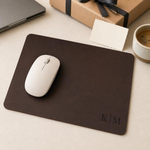 May include: A rectangular dark brown leather mouse pad with rounded corners. The bottom right corner features a debossed monogram with the letters K and M separated by a vertical line. A smooth, off-white wireless computer mouse rests on the surface.
