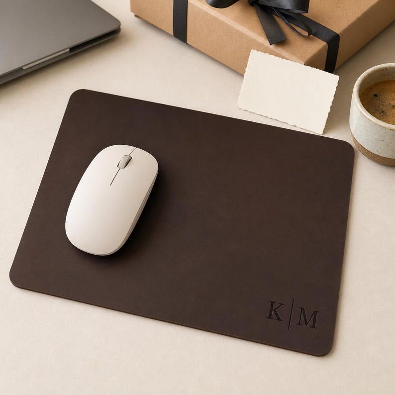 May include: A rectangular dark brown leather mouse pad with rounded corners. The bottom right corner features a debossed monogram with the letters K and M separated by a vertical line. A smooth, off-white wireless computer mouse rests on the surface.