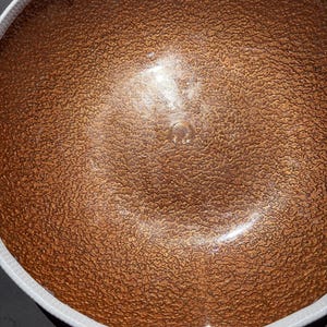 May include: A round bowl featuring a textured, metallic copper-colored interior surface with a distinct crinkled or hammered appearance. The bowl has a smooth, plain white exterior rim that contrasts with the deep, reflective bronze-toned center.