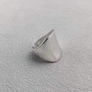 Könnte beinhalten: Ein silberfarbener Ring mit einer breiten, geschwungenen Oberfläche. Der Ring hat eine glatte, polierte Oberfläche und ein schlichtes, modernes Design. Der Ring ist wahrscheinlich aus Sterlingsilber oder einem ähnlichen Metall gefertigt.