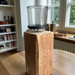 Könnte beinhalten: Ein klarer Glas-Kerzenhalter sitzt auf einem dunklen Metallsockel, der an einem rustikalen, rechteckigen Holzblock befestigt ist. Das Holz hat ein natürliches, verwittertes Aussehen. Der Kerzenhalter steht auf einem runden Holztisch.