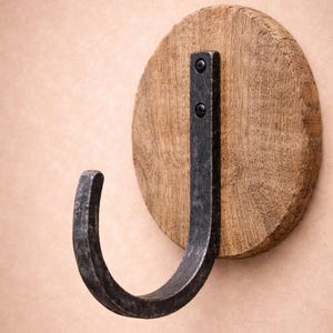 May include: A rustic wall hook featuring a dark metal hook attached to a round, light brown wooden base. The hook is curved and has a distressed finish, while the wood grain is visible on the base. Two screws secure the metal to the wood.