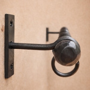 Puede incluir: Un toallero de hierro forjado con acabado oscuro. El soporte de pared tiene una base rectangular con orificios para tornillos. El anillo está unido a una barra horizontal y una esfera texturizada.