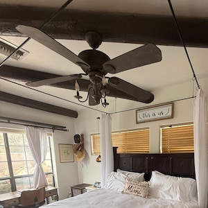 May include: A bedroom with a dark wood bed frame, white bedding, and a decorative pillow that says "Mimi". A ceiling fan with dark blades hangs above the bed. The room has white curtains and a sign that says "This is us".