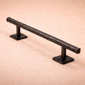 Puede incluir: Una barra de metal oscuro, hecha a mano, con una superficie texturizada, soportada por dos bases cuadradas. La barra mide aproximadamente 30 cm de largo, ideal para un tirador de armario. El herraje tiene una estética industrial rústica.