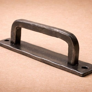 May include: A dark grey, hand-forged metal handle with a rectangular base and a curved grip. The handle has a rustic, textured appearance and two holes for mounting. The hardware is likely for a cabinet or door.