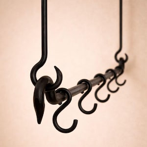Puede incluir: Perchero de metal negro con múltiples ganchos en forma de S. El perchero está suspendido de una barra vertical y tiene un aspecto rústico y artesanal. Los ganchos están espaciados uniformemente a lo largo de una barra horizontal.