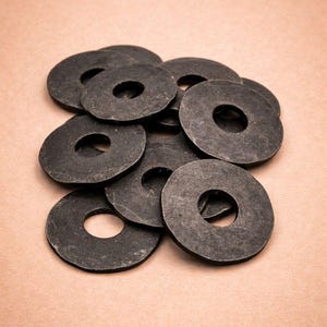 Op de afbeelding: Een verzameling donkergrijze, cirkelvormige ringen met een centraal gat. Ze liggen op een lichtbruin oppervlak. De afbeelding is een close-up, die de textuur van rubber of een vergelijkbaar materiaal laat zien. De ringen zijn waarschijnlijk duurzaam.