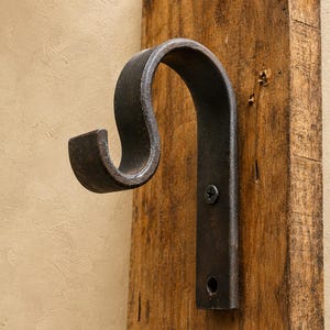 Puede incluir: Un gancho de metal gris oscuro curvado con una placa de montaje plana fijada a una tabla de madera vertical. El gancho presenta una silueta en forma de S y está asegurado a la madera con un tornillo negro visible en un orificio perforado.